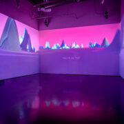 Three walls in a gallery with projections on them. The projections depict a pink sky and abstract purple mountains.