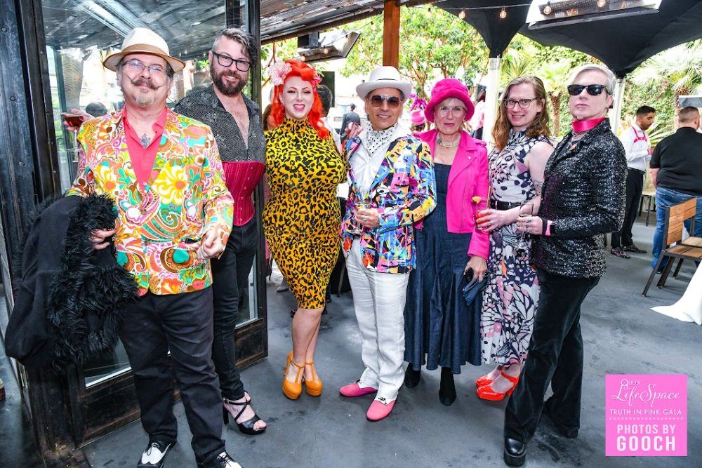 a group of 7 queers wearing colorful, flamboyant outfits posing together