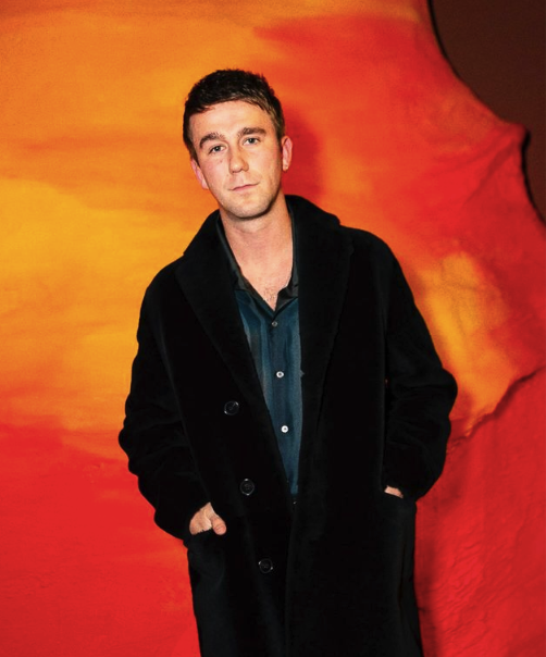 SG Lewis poses with his hands in the pockets of a long black over coat