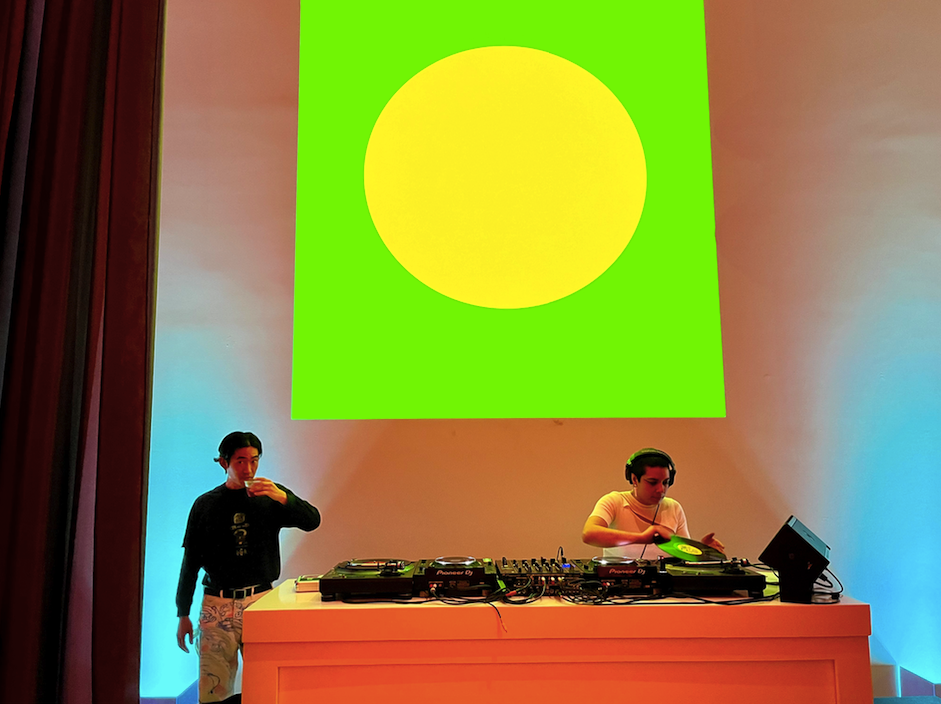 a set of DJs entertain the crowd with a large circular pop art piece behind them