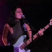 Maya, a woman with black hair and a member of Denver based band Soph and Austin, is holding a light green bass guitar and singing into a microphone.