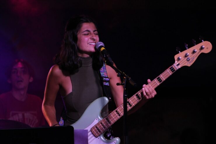 Maya, a woman with black hair and a member of Denver based band Soph and Austin, is holding a light green bass guitar and singing into a microphone.