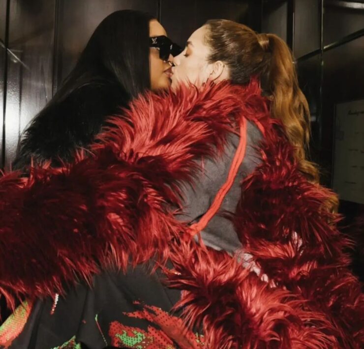 First Daughter of Cameroon, Brenda Biya, a black woman with black straight hair and sunglasses, kisses Brazilian model Layyons Valenca, a white woman with light brown hair in a high ponytail wearing a black and red feathered outfit.