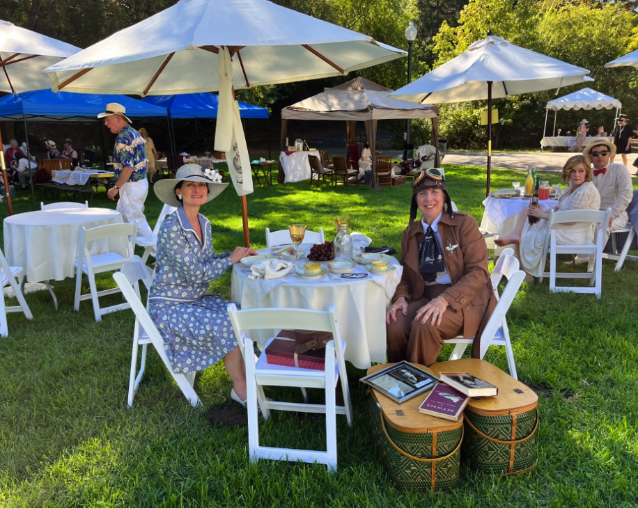 a woman dressed like Amelia Airhart and friend sit at tables beneath umbrellas