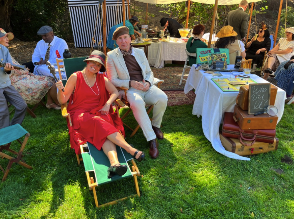 a couple poses on lawn chairs near a set up of vintage board games like CLUE