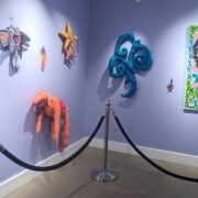 Image of artwork by Starr Sprite hung on lavender painted walls. The image shows two walls, with canned lights shining down on the art. There are eight pieces of art of various sizes on the wall. This artwork is mostly sewn fabric featuring imaginative sewn characters.