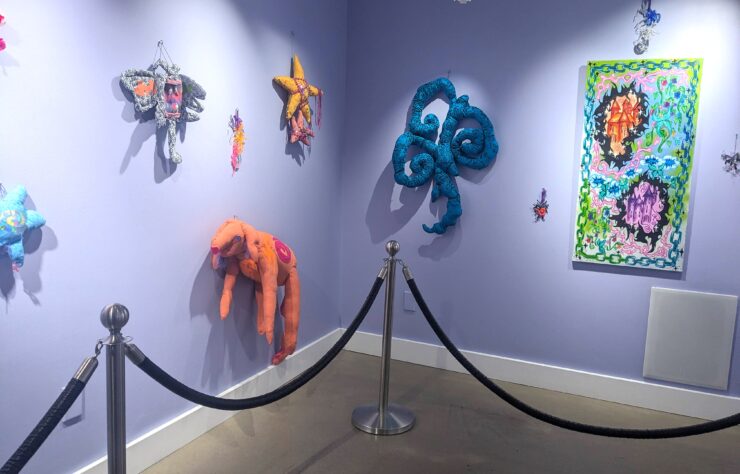 Image of artwork by Starr Sprite hung on lavender painted walls. The image shows two walls, with canned lights shining down on the art. There are eight pieces of art of various sizes on the wall. This artwork is mostly sewn fabric featuring imaginative sewn characters.