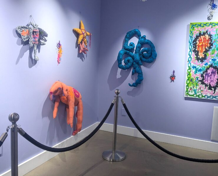 Image of artwork by Starr Sprite hung on lavender painted walls. The image shows two walls, with canned lights shining down on the art. There are eight pieces of art of various sizes on the wall. This artwork is mostly sewn fabric featuring imaginative sewn characters.