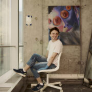 This image shows a picture of the artist featured in this interview, wearing a white t-shirt and jeans looking off in the distance. The artist is sitting in their studio on a white rolling office chair in front of a colorful painting hung on a concrete wall.