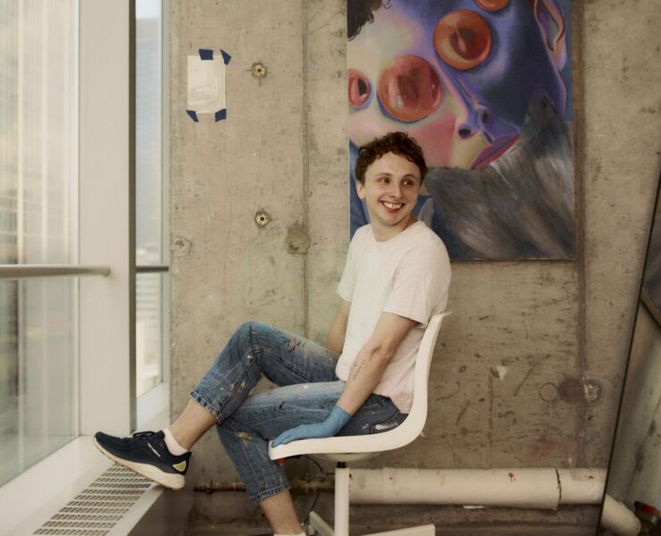 This image shows a picture of the artist featured in this interview, wearing a white t-shirt and jeans looking off in the distance. The artist is sitting in their studio on a white rolling office chair in front of a colorful painting hung on a concrete wall.