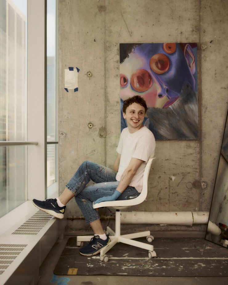 This image shows a picture of the artist featured in this interview, wearing a white t-shirt and jeans looking off in the distance. The artist is sitting in their studio on a white rolling office chair in front of a colorful painting hung on a concrete wall.