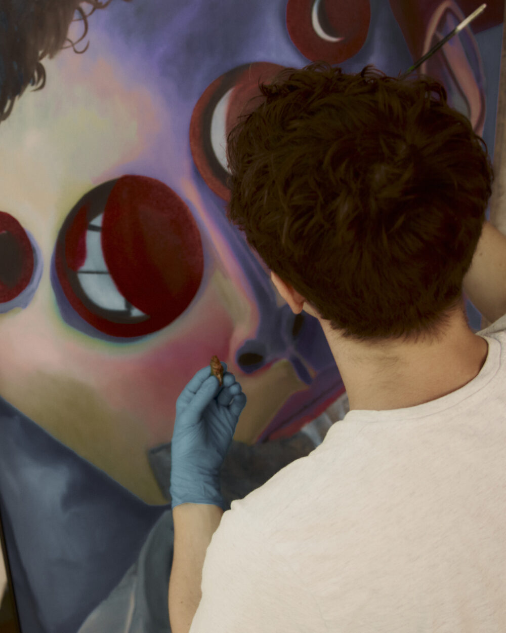 Image of artist Eli Hill painting in their studio. It is shot from above and shows the back of the artist's head and shoulders with a white t-shirt. He has on blue rubber gloves and is painting an oil painting with purple circles.