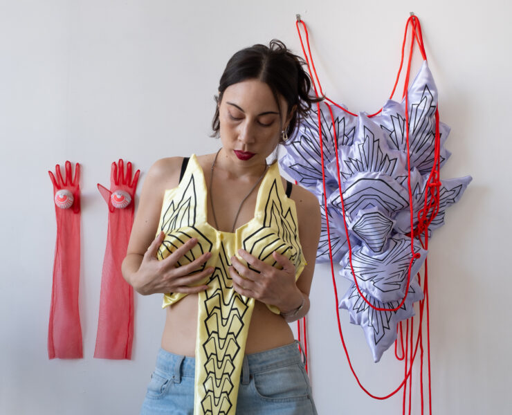 A portrait photograph of Chanel Matsunami holding up a yellow silk sculpture as if it were clothing. They are standing in front of two long pink gloves hung on a white wall next to a pink and purple silk wall sculpture