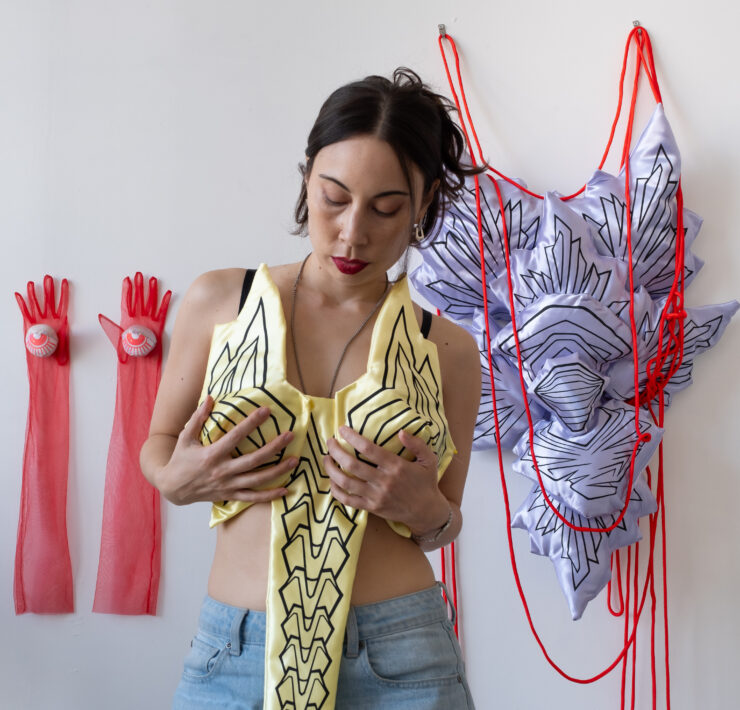 A portrait photograph of Chanel Matsunami holding up a yellow silk sculpture as if it were clothing. They are standing in front of two long pink gloves hung on a white wall next to a pink and purple silk wall sculpture