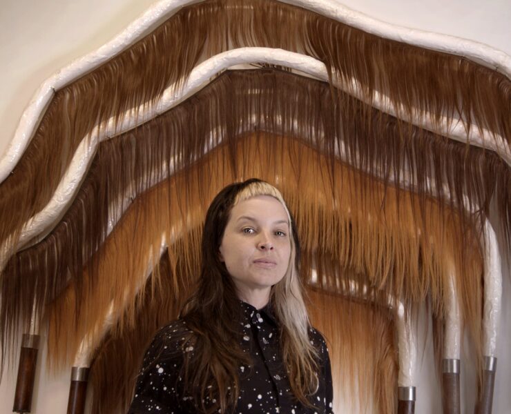 An image of an artist facing forward looking at the audience. They are in a dark button up shirt with small light dots. They are looking at the audience. They are standing in front of five sewn arches with brown and camel colored fake hair hanging from each arch. The arches are about eight feet tall and wide.