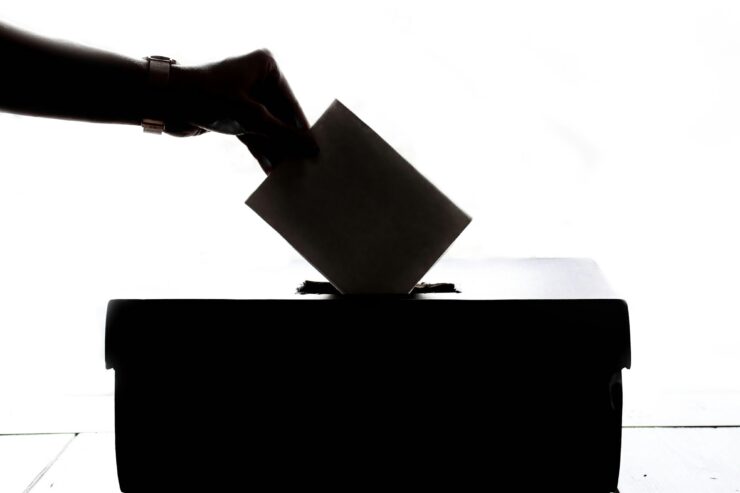 Exit Polls Show 86% of Queer Voters Elected Kamala Harris: On a white backdrop a hand placing an envelope into a box is in silhouette