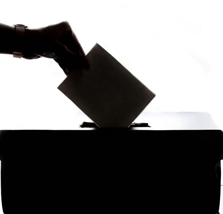 Exit Polls Show 86% of Queer Voters Elected Kamala Harris: On a white backdrop a hand placing an envelope into a box is in silhouette