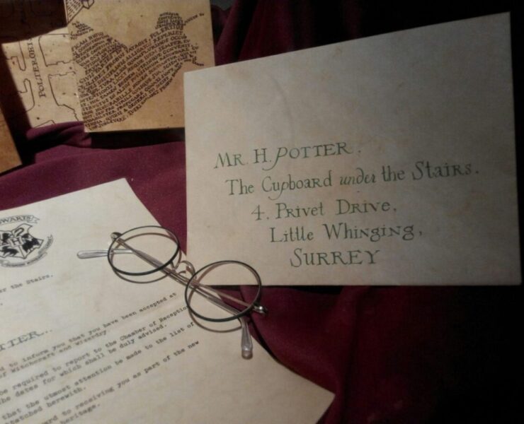 HBO Defends J.K. Rowling’s Involvement In Harry Potter Series: An envelope from hogwarts to harry potter with his name and address. A pair of circle glasses sits next to in on top of the letter accepting him into the school. All is sitting on top of a dark red table cloth