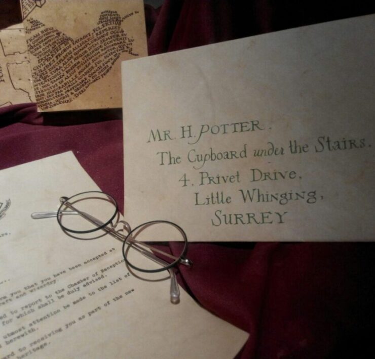HBO Defends J.K. Rowling’s Involvement In Harry Potter Series: An envelope from hogwarts to harry potter with his name and address. A pair of circle glasses sits next to in on top of the letter accepting him into the school. All is sitting on top of a dark red table cloth