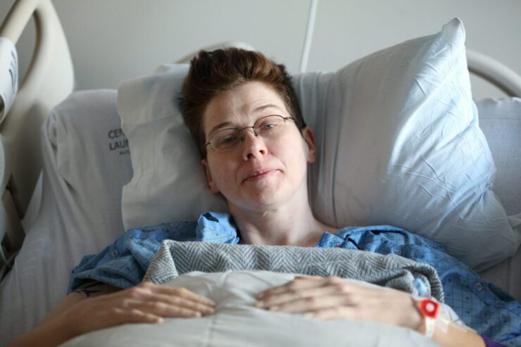 Denver Health Continues to Provide LGBTQ+ Health Services: A queer person with short brown hair and glasses sits in a hospital bed looking at the camera