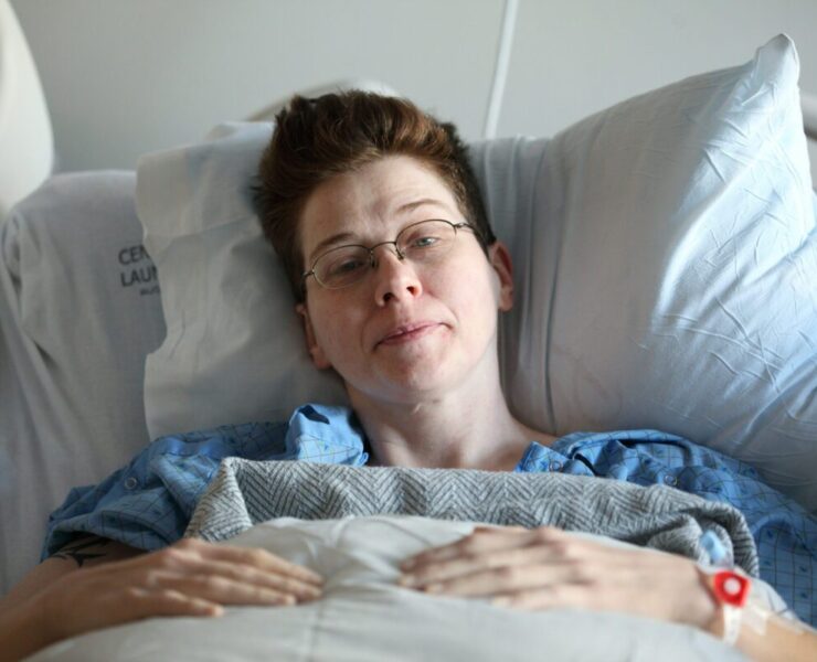 Denver Health Continues to Provide LGBTQ+ Health Services: A queer person with short brown hair and glasses sits in a hospital bed looking at the camera