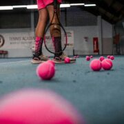 A trans girl stands on a tennis court surrounded by pink tennis balls.