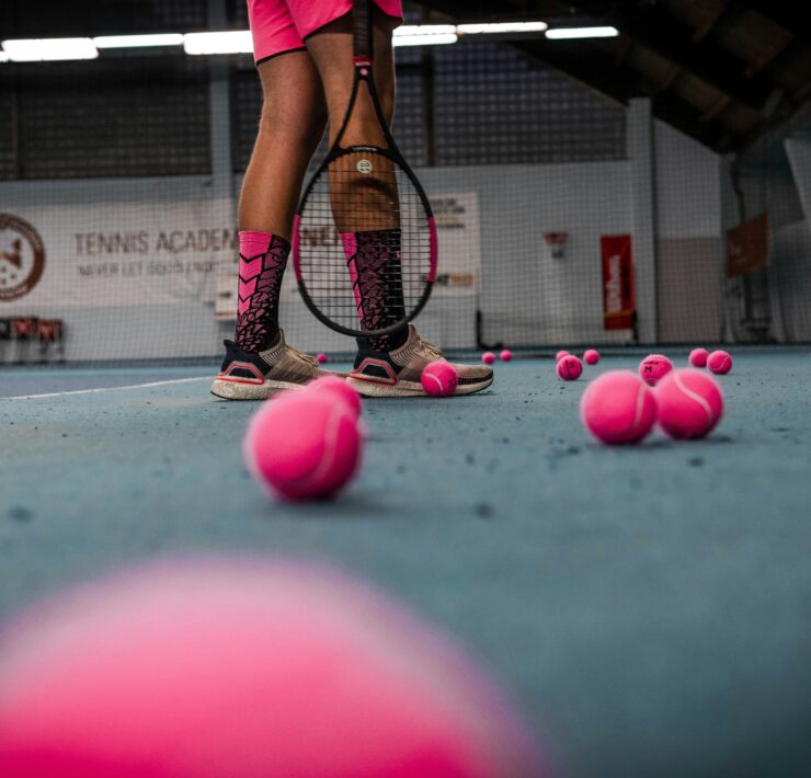 A trans girl stands on a tennis court surrounded by pink tennis balls.