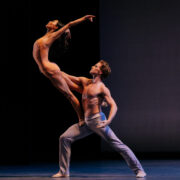 two minimally dressed dancers strike a complicated lift pose with the female dancer balanced on the male dancer's thigh with chest arched forward and arms stretched in the air