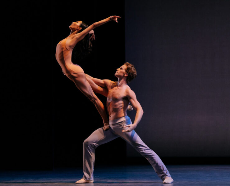 two minimally dressed dancers strike a complicated lift pose with the female dancer balanced on the male dancer's thigh with chest arched forward and arms stretched in the air