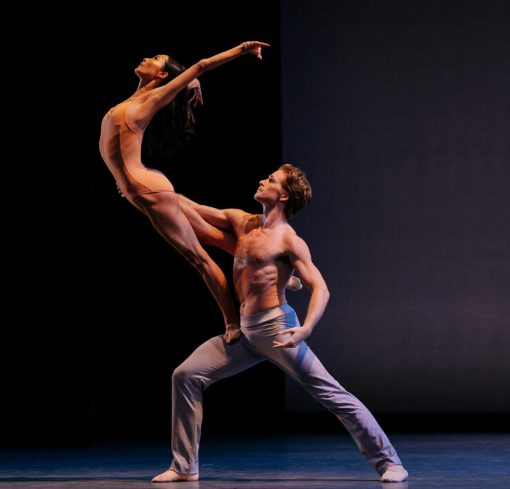 two minimally dressed dancers strike a complicated lift pose with the female dancer balanced on the male dancer's thigh with chest arched forward and arms stretched in the air