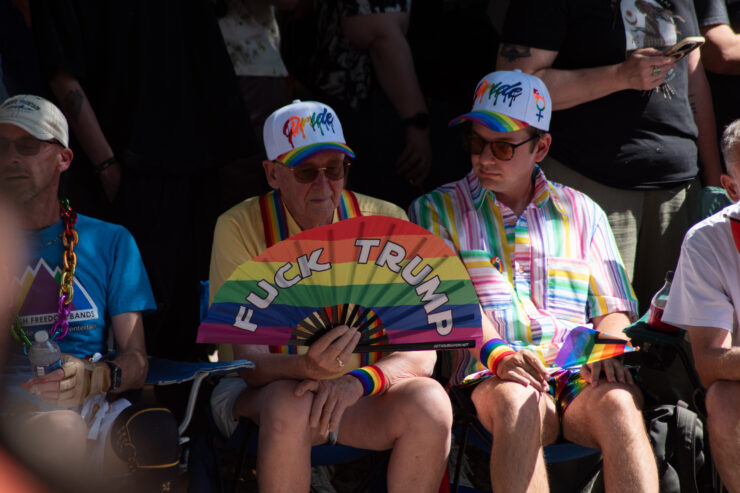 A closeup on the crowd watching the parade. One individual holds a rainbow fan that says Fuck Trump on it.