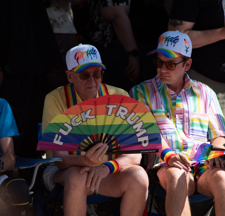 A closeup on the crowd watching the parade. One individual holds a rainbow fan that says Fuck Trump on it.