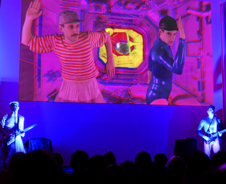The two artists in Princess Collaborative performing on a stage. They stand holding stringed instruments facing an audience on the left and right sides of the stage lit with a light blue light. Behind them a large projection of a music video featuring the artists is playing. The video features a magenta hue.