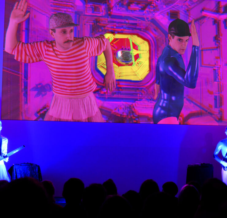 The two artists in Princess Collaborative performing on a stage. They stand holding stringed instruments facing an audience on the left and right sides of the stage lit with a light blue light. Behind them a large projection of a music video featuring the artists is playing. The video features a magenta hue.