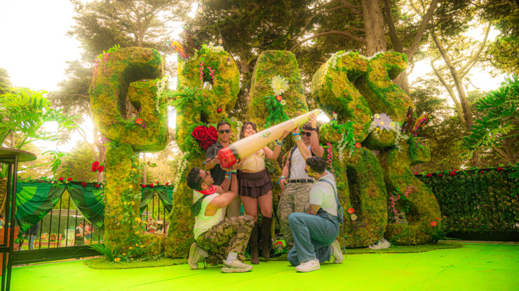 several festival goers posse in front of the large topiary grass lands sign with an inflatable joint