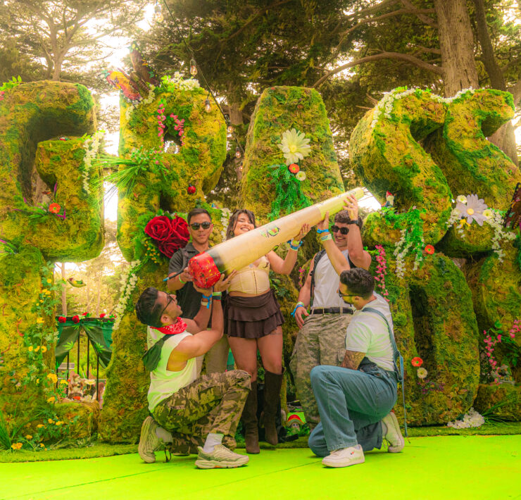 several festival goers posse in front of the large topiary grass lands sign with an inflatable joint
